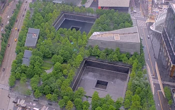 Aerial view of the World Trade Center Memorial in New York City, showing the two large reflecting pools surrounded by trees and bronze name panels, symbolizing remembrance and resilience.