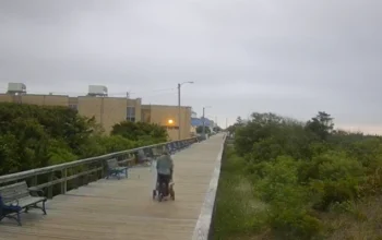 Live view of Avalon New Jersey Boardwalk showing a peaceful wooden path along the shoreline with ocean waves, sandy beach, and clear skies in the background.