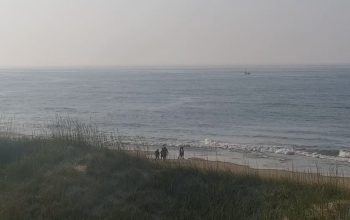 Frisco Woods Campground live cam view of the beach with ocean waves, sandy shoreline, and distant dunes under a clear sky