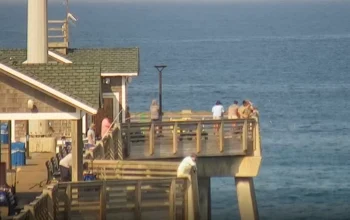 Live view from Jennette’s Pier Cam showing the oceanfront pier stretching into the Atlantic, with sandy Nags Head beach, gentle waves, and clear blue skies in the background.
