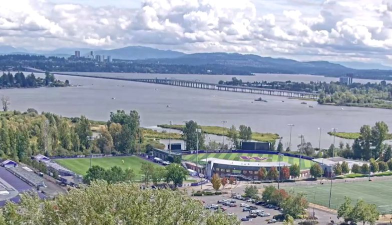 University of Washington live webcam view overlooking Lake Washington with calm blue water, tree-lined shores, and Seattle skyline in the distance.