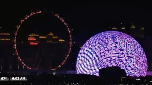 Las Vegas Sphere at night glowing with vibrant colors, with the High Roller Ferris wheel and Strip hotels in the distance