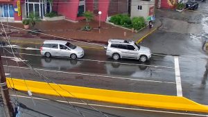 Live webcam view of Barbican neighborhood in Kingston, Jamaica, showing busy streets with cars, pedestrians, colorful shops, and tropical greenery under bright Caribbean sunlight.