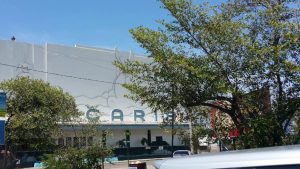 Live webcam view of Carib 5 cinema facing Cross Roads in Kingston, Jamaica, showing bustling streets with taxis, pedestrians, and the brightly lit theater surrounded by city lights and tropical evening energy.