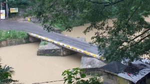 Live webcam view of Flat Bridge crossing the Rio Cobre River in St. Catherine, Jamaica, showing vehicles carefully driving over the narrow, rail-less bridge surrounded by lush green hills and flowing water below.