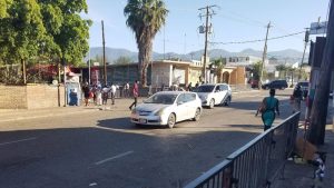 Live webcam view of Half Way Tree in Kingston, Jamaica, showing the historic Clock Tower surrounded by bustling traffic, street vendors, and pedestrians moving through the busy city center under bright tropical skies.