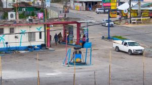 Live webcam view of Little London in Westmoreland, Jamaica, showing a quiet rural town with passing cars, colorful roadside shops, and locals walking under bright tropical sunlight surrounded by lush green vegetation.