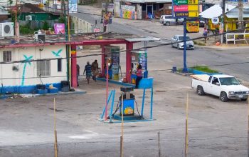 Live webcam view of Little London in Westmoreland, Jamaica, showing a quiet rural town with passing cars, colorful roadside shops, and locals walking under bright tropical sunlight surrounded by lush green vegetation.