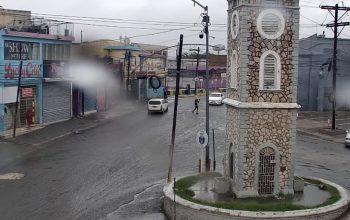 Live webcam view of downtown May Pen, Jamaica, showing busy streets with cars, market vendors, and pedestrians moving through the town center near the Rio Minho River under bright tropical skies.